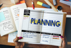 Manos femeninas sostienen una agenda física, sobre un escritorio donde hay otros objetos utilizados para organizar la productividad personal.