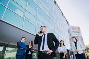 Un grupo de personas interactúan y una habla por teléfono, de fondo se ve la fachada de un edificio corporativo, para ilustrar los entornos de las grandes empresas.