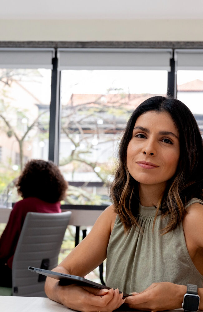 Una mujer posa en una oficina con amplias ventanas y colegas trabajando en segundo plano. El ambiente es profesional y moderno, dentro de una empresa mediana.