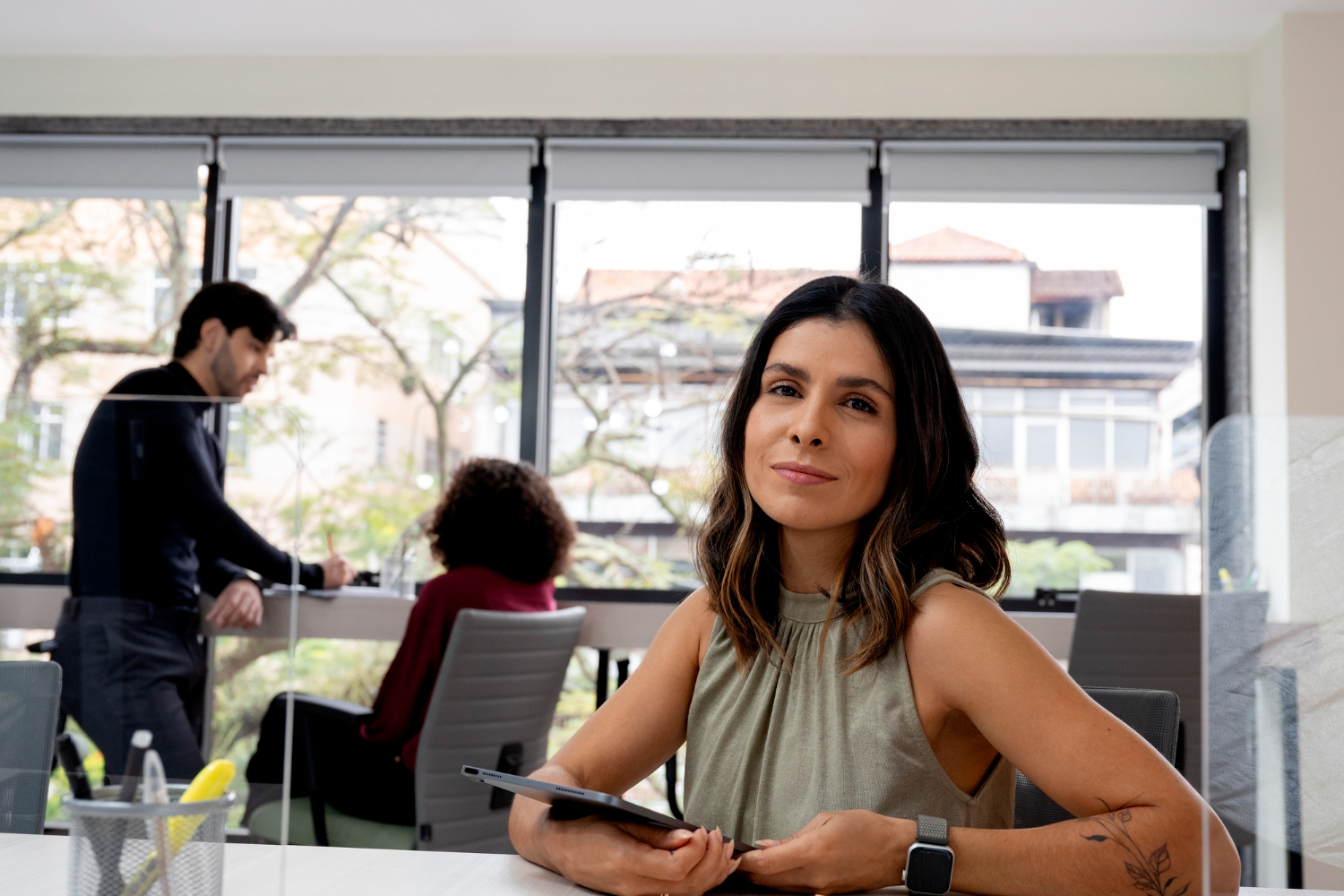 Una mujer posa en una oficina con amplias ventanas y colegas trabajando en segundo plano. El ambiente es profesional y moderno, dentro de una empresa mediana.