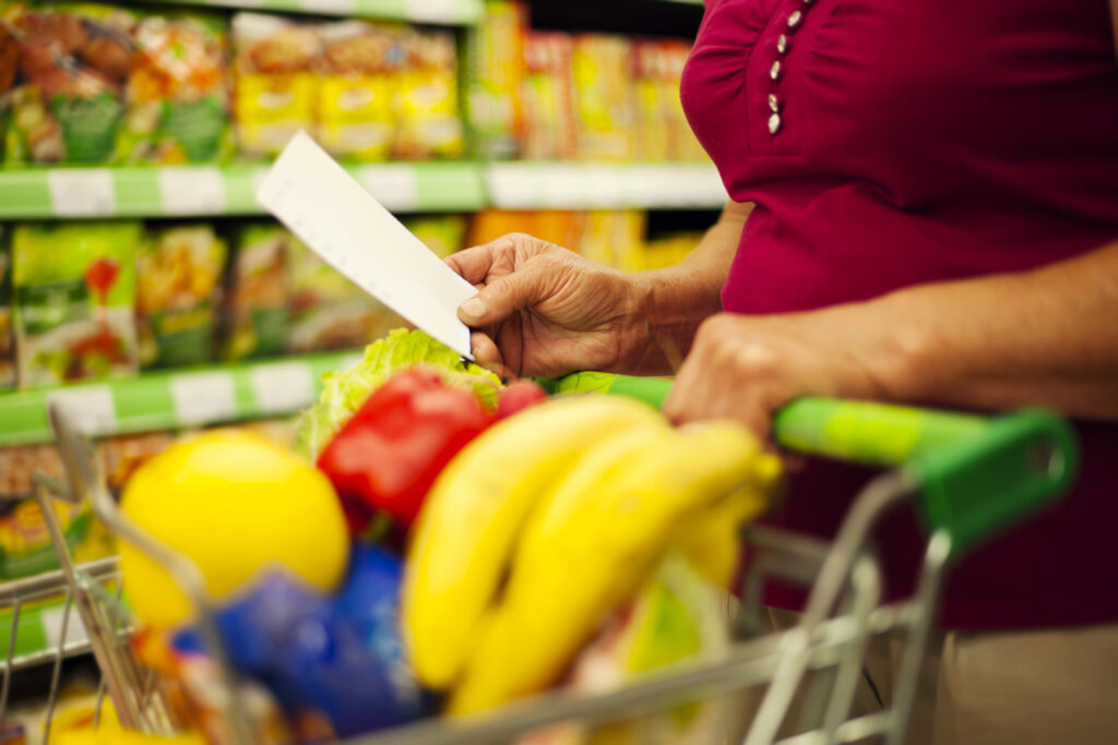 Una persona con blusa roja revisa una lista de compras mientras empuja un carrito lleno de frutas y verduras frescas, dentro de un comercio local.