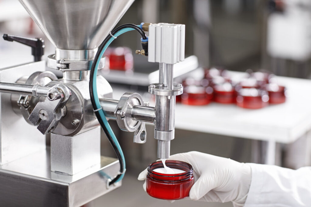 Una mano enguantada sostiene un frasco rojo mientras una máquina industrial dispensa un producto en un proceso de producción de una empresa.