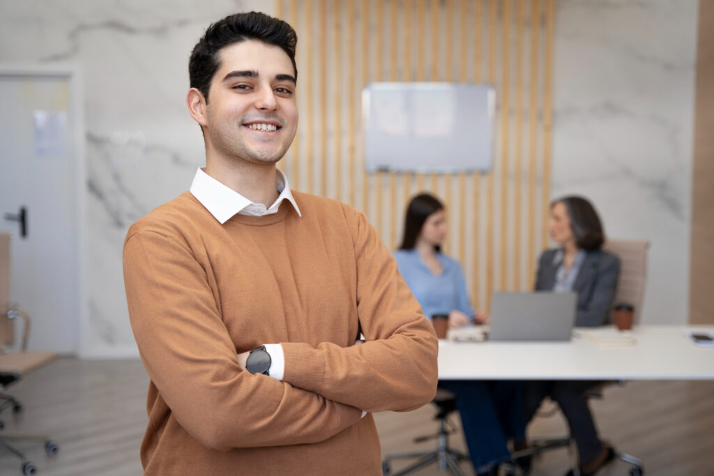 Un hombre joven sonriente posa en una oficina moderna de una empresa mediana en México. Al fondo, dos mujeres trabajan en un escritorio.