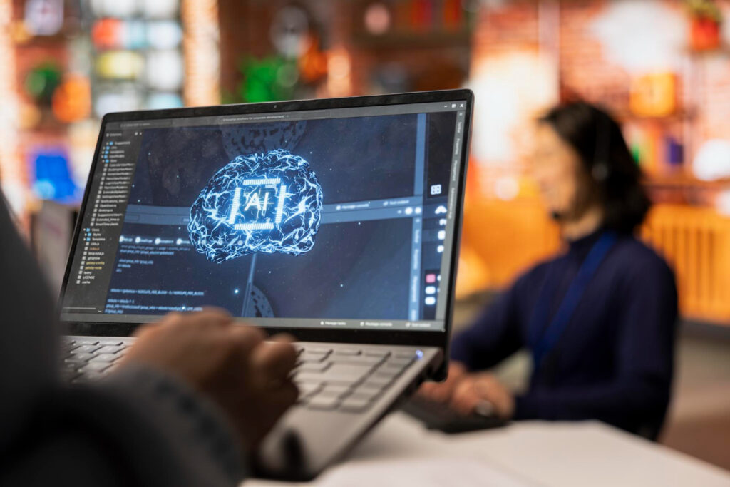 Persona trabajando en una computadora portátil que tiene en la pantalla un esquema del cerebro con la frase AI en el centro. La imagen ilustra las aplicaciones de la Inteligencia Artificial en las empresas.