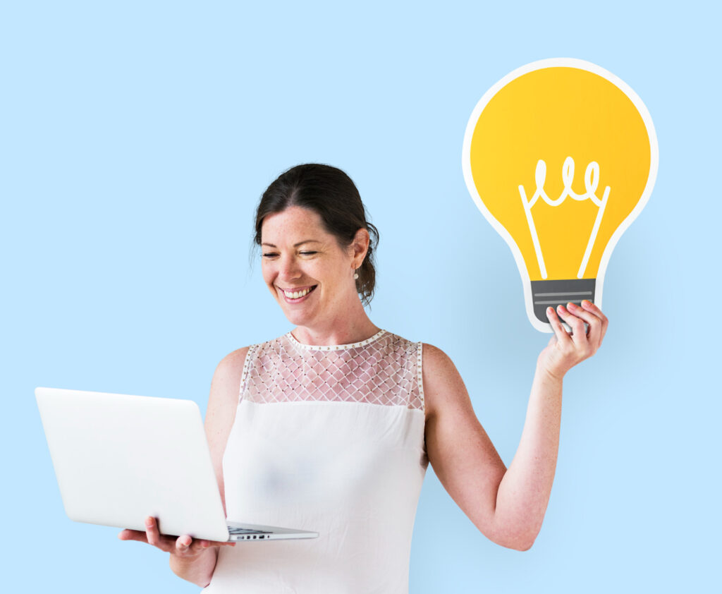 Una mujer sonriente, de mediana edad, sostiene una computadora portátil con una mano y una gran bombilla de idea simboliza su mentalidad emprendedora.
