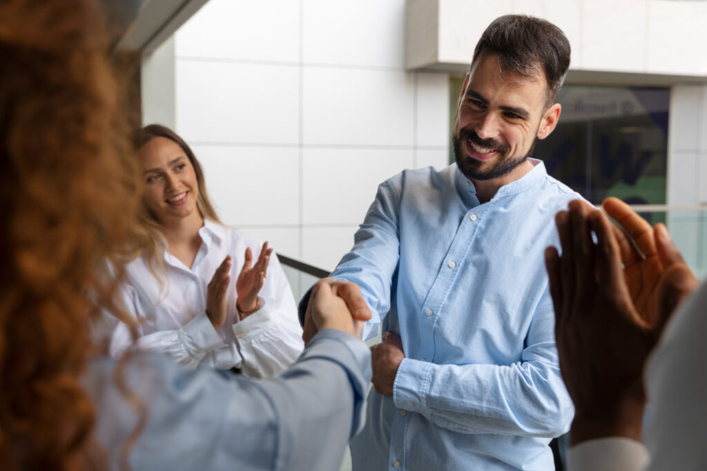 Como muestra de su liderazgo, un hombre ofrece su reconocimiento a una colaboradora, con un apretón de manos, mientras otras personas aplauden.