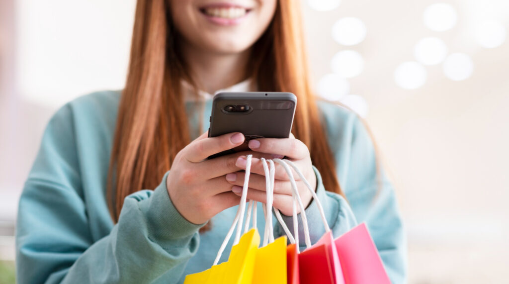 Vista de una mujer que sostiene un teléfono celular con el que está haciendo compras en línea. También sostiene bolsas de compra.