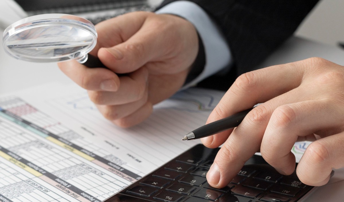Manos masculinas sostienen una lupa que permite revisar datos contenidos en hojas impresas sobre un escritorio. La otra sostiene una pluma y realiza operaciones en una calculadora.