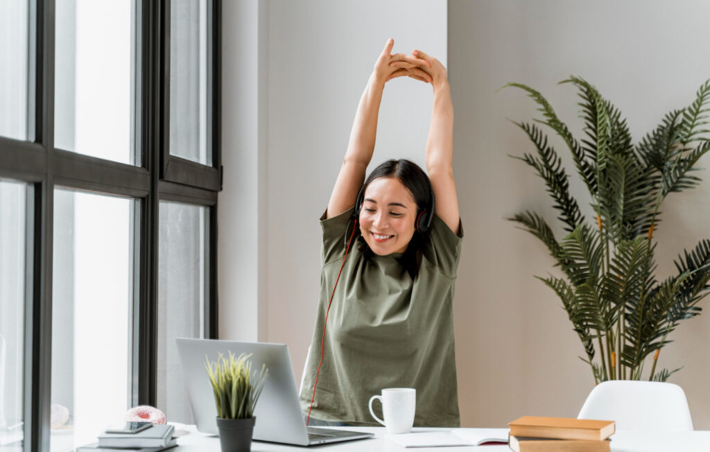 Mujer con audífonos, sentada frente a una computadora portátil, sonríe y se estira mientras mira la computadora, en señal de bienestar laboral.