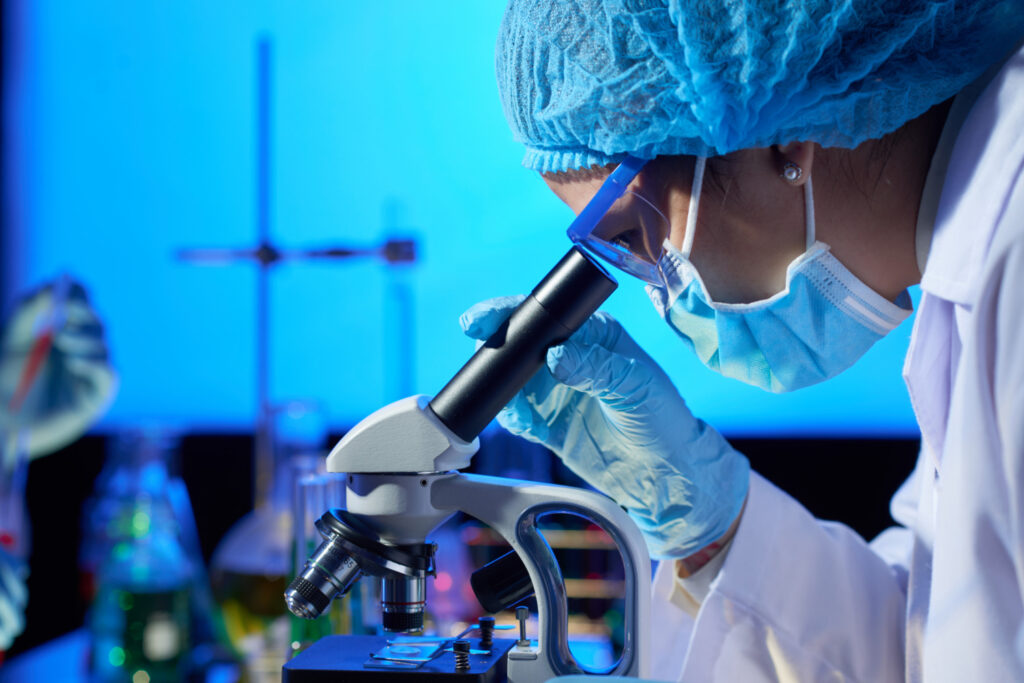 Una mujer que porta bata, guantes y cofia manipula un microscopio en un laboratorio. La imagen ilustra una biocientífica.