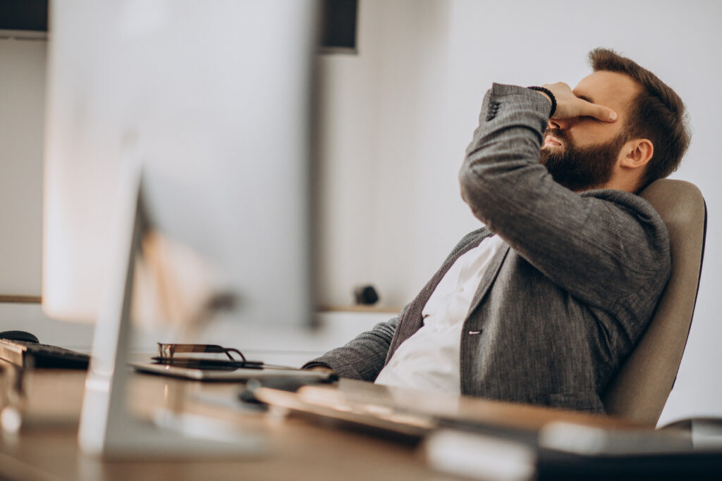 Hombre sentado en un escritorio se cubre los ojos en señal de agotamiento mental o burnout.