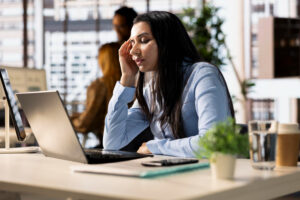 Mujer sentada en un escritorio, frente a una computadora, muestra señales de agotamiento. De fondo se aprecia un ambiente de oficina.