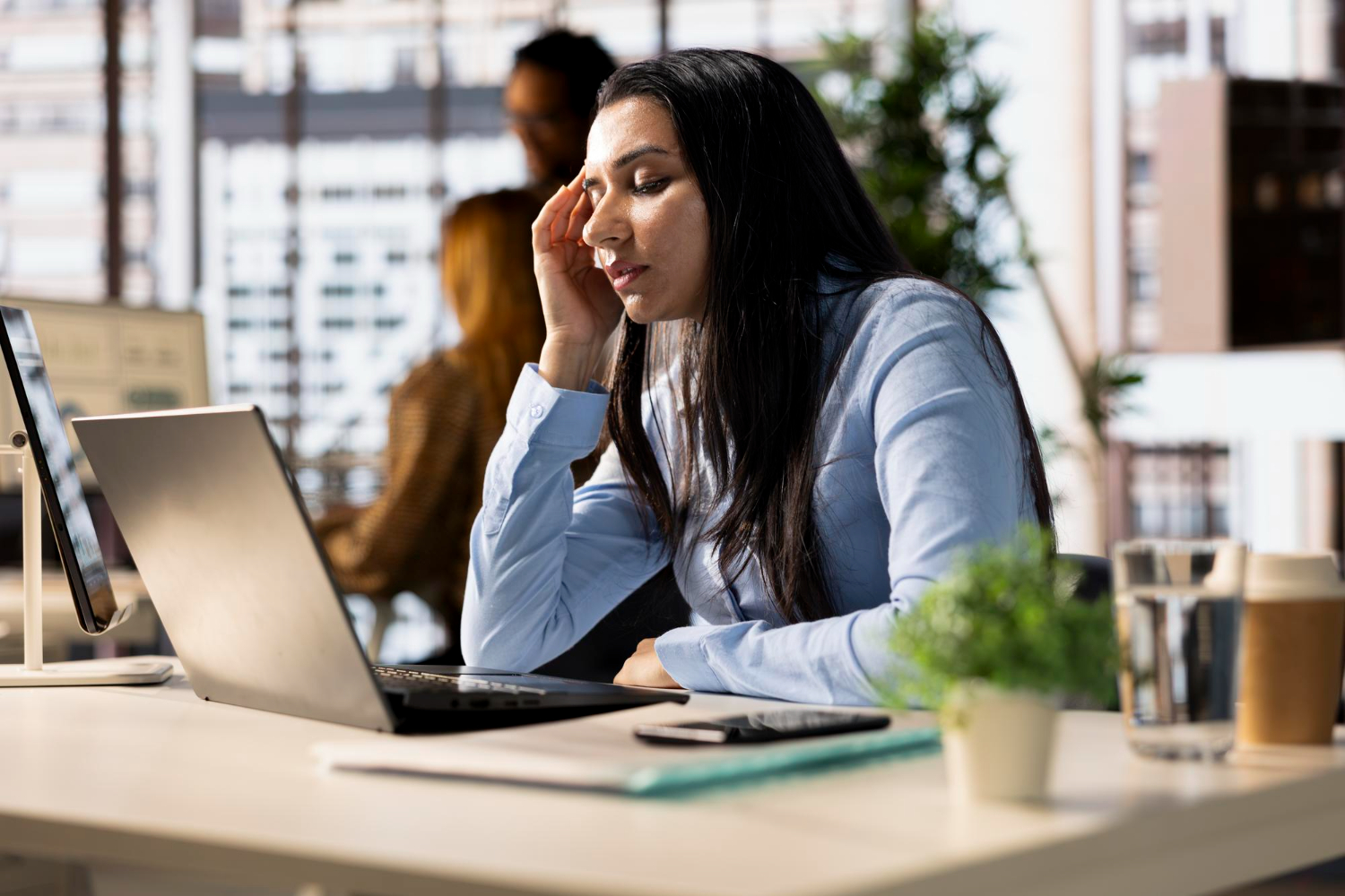 Mujer sentada en un escritorio, frente a una computadora, muestra señales de agotamiento. De fondo se aprecia un ambiente de oficina.