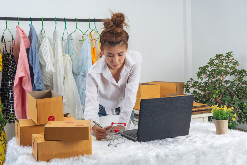 Una mujer realiza anotaciones. En su escritorio hay una computadora, cajas de paquetería y un carrito de compras. De fondo tiene un inventario de ropa.