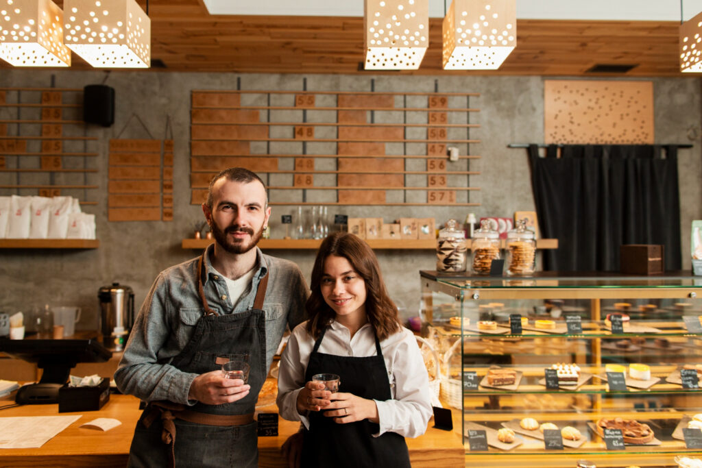 Un hombre y una mujer miran a la cámara; de fondo se aprecia una barra y vitrina de una cafetería. La imagen remite a dos emprendedores.