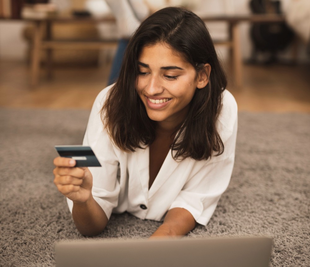 Una mujer recostada en el piso, frente una laptop, sonríe mientras mira una tarjeta bancaria. La imagen ilustra una compra en línea.
