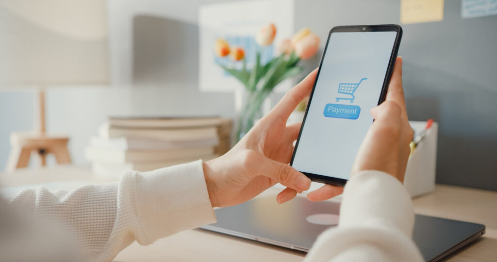 Unas manos sostienen una tableta electrónica que es usada para realizar compras en línea, de fondo se ve un escritorio donde hay una laptop, flores y otros elementos.