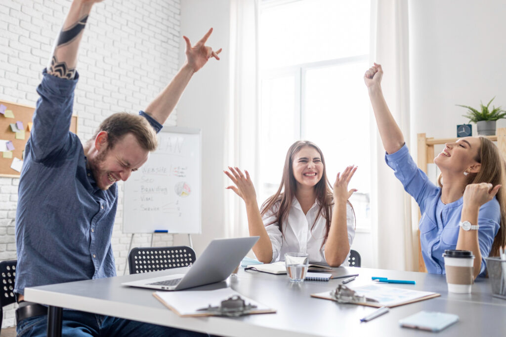 Dos mujeres y un hombre, sentados en una sala de juntas de una oficina, celebran con gestos de alegría y sus manos levantadas.