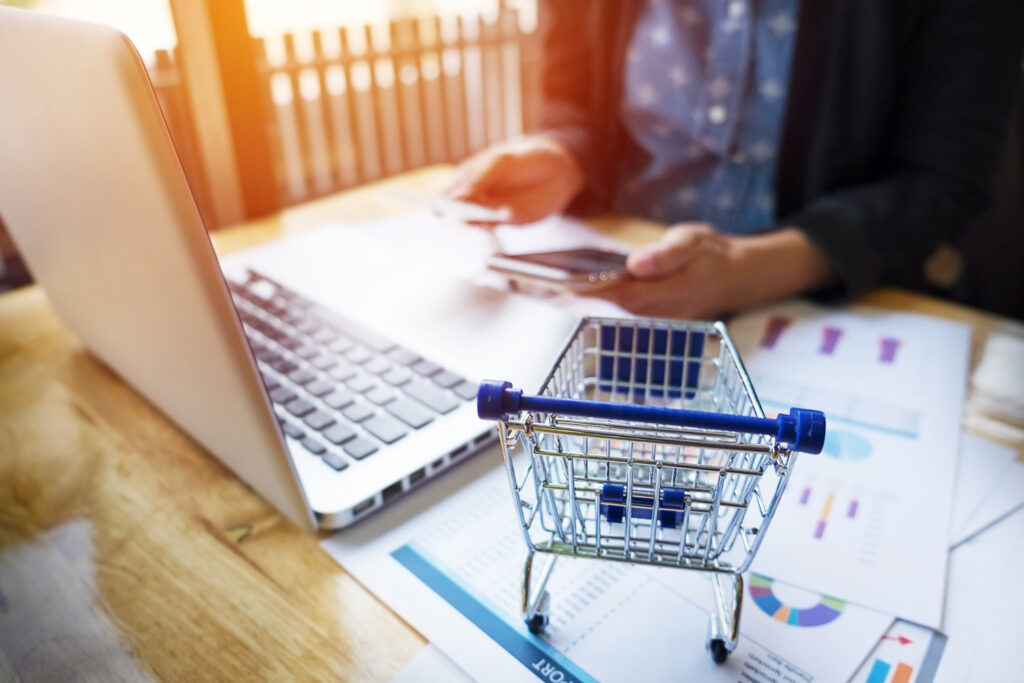 Una persona sentada frente a una computadora, usa un teléfono celular y una tarjeta bancaria. Sobre su escritorio hay un carrito de compras y gráficas impresas.