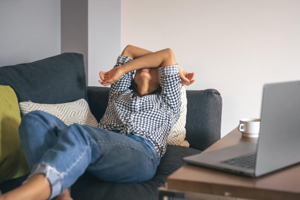 Mujer recostada en un sofá se cubre la cara con los brazos. En la mesa de la sala se ve una computadora portátil.