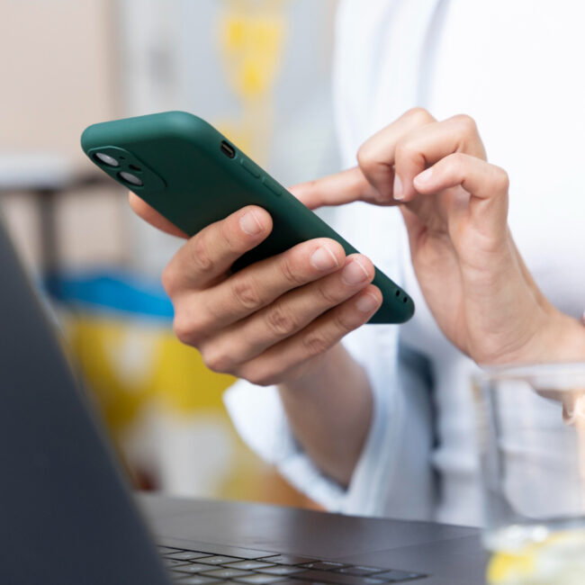 Una persona con un teléfono celular en la mano envía mensajes. Enfrente tiene una computadora portátil.