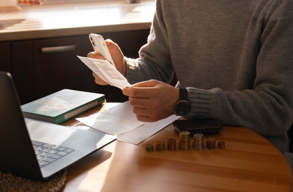 Vista de las manos de un hombre que revisa cuentas, sentado frente a su computadora. Sostiene un teléfono celular y un comprobante de pago.
