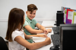 Niños sentados frente a dos computadoras sonríen entre ellos. La imagen ilustra el aprendizaje de programación en la infancia.
