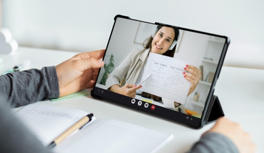 Una persona sostiene una tableta frente a sí, donde está la transmisión de una persona que expone un tema. La imagen ilustra la capacitación en línea.