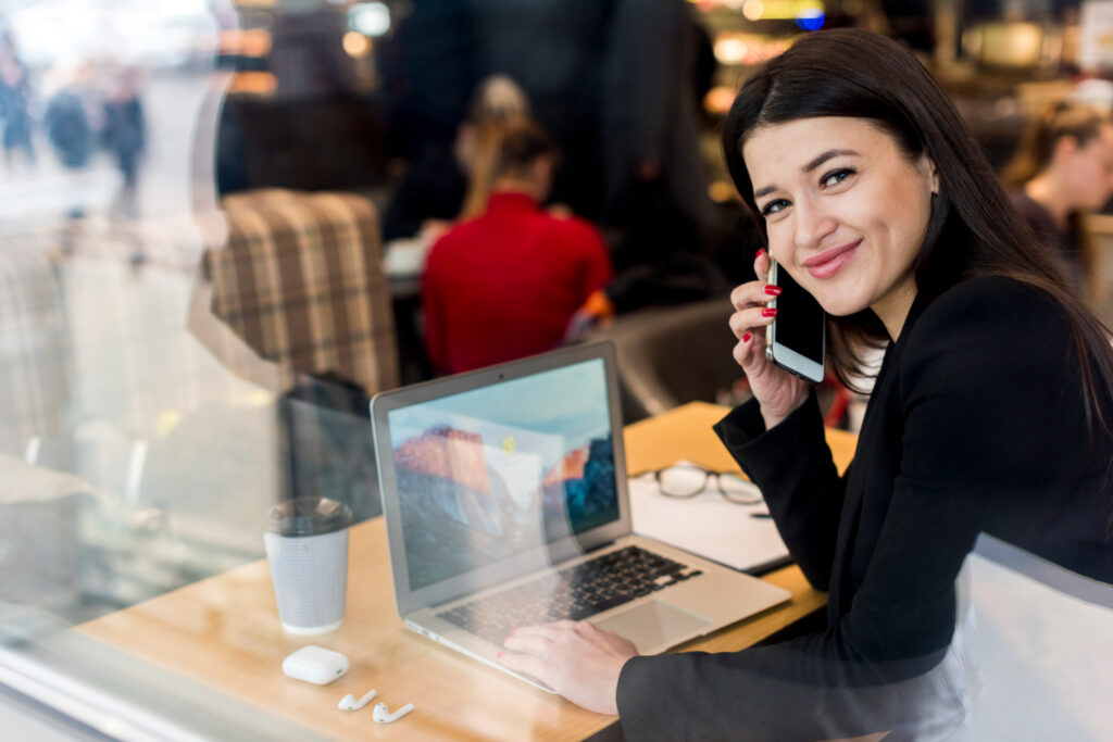 Una mujer sentada en un escritorio, frente a una computadora, sonríe hacia la cámara, mientras sostiene un teléfono. De fondo se ven otras personas en un ambiente de trabajo.