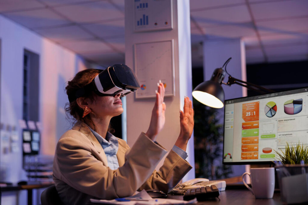 Mujer, en una oficina, sentada en un escritorio, usa unos lentes de realidad virtual. A un lado de ella hay una computadora con gráficas. 