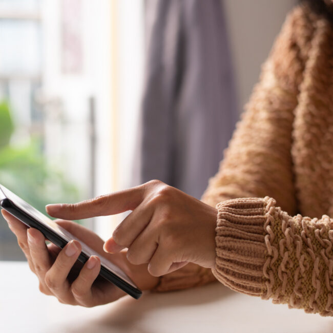 Primer plano de las manos de una persona sosteniendo y utilizando un teléfono inteligente. La persona viste un suéter de punto color marrón texturizado y está sentada frente a una mesa blanca.