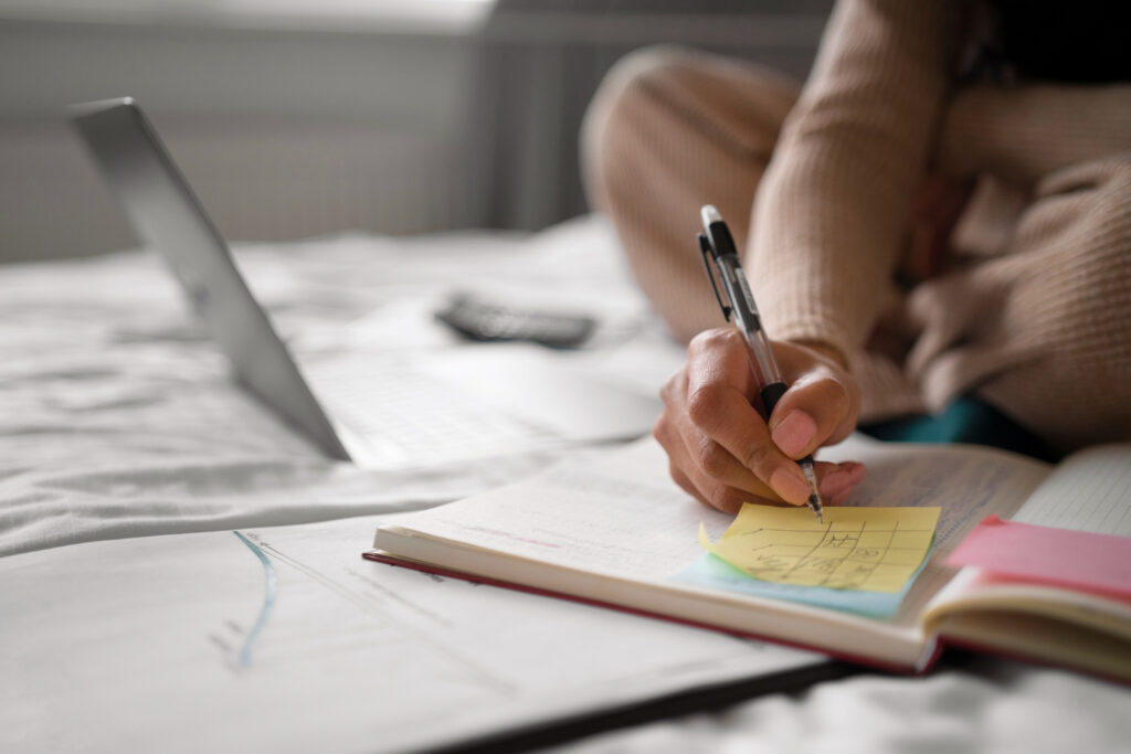 Una persona, sentada en una cama, escribe sus actividades en una agenda de papel. A un lado se observa una computadora portátil.