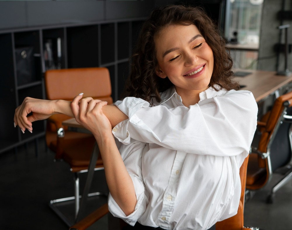Mujer, sentada en un espacio de trabajo, esboza una sonrisa mientras hace un movimiento de estiramiento con un brazo, para ilustrar los periodos de descanso de la técnica Pomodoro. 