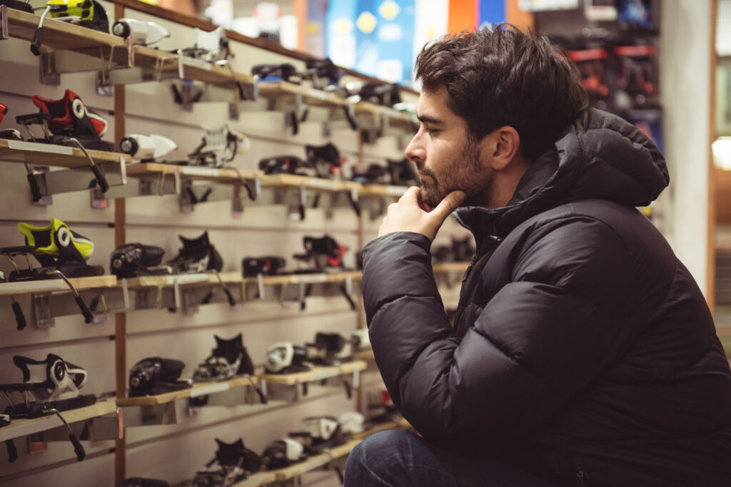 Un hombre con barba y chamarra de esquí mira pensativo, un estante lleno de accesorios para esquiar, en una tienda deportiva. La imagen ilustra intereses de los consumidores.