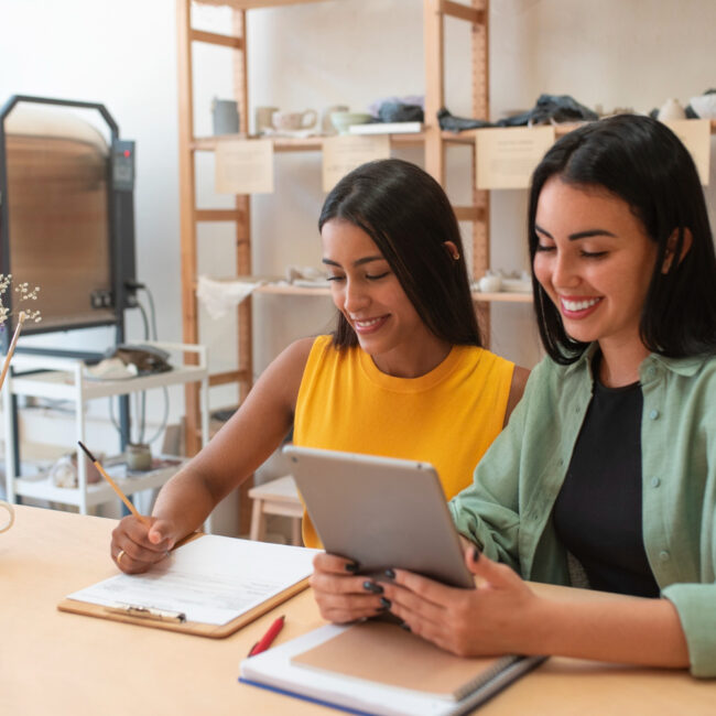 Dos mujeres sentadas en un escritorio trabajan juntas. De fondo se ve un anaquel. La imagen representa a dos emprendedoras.