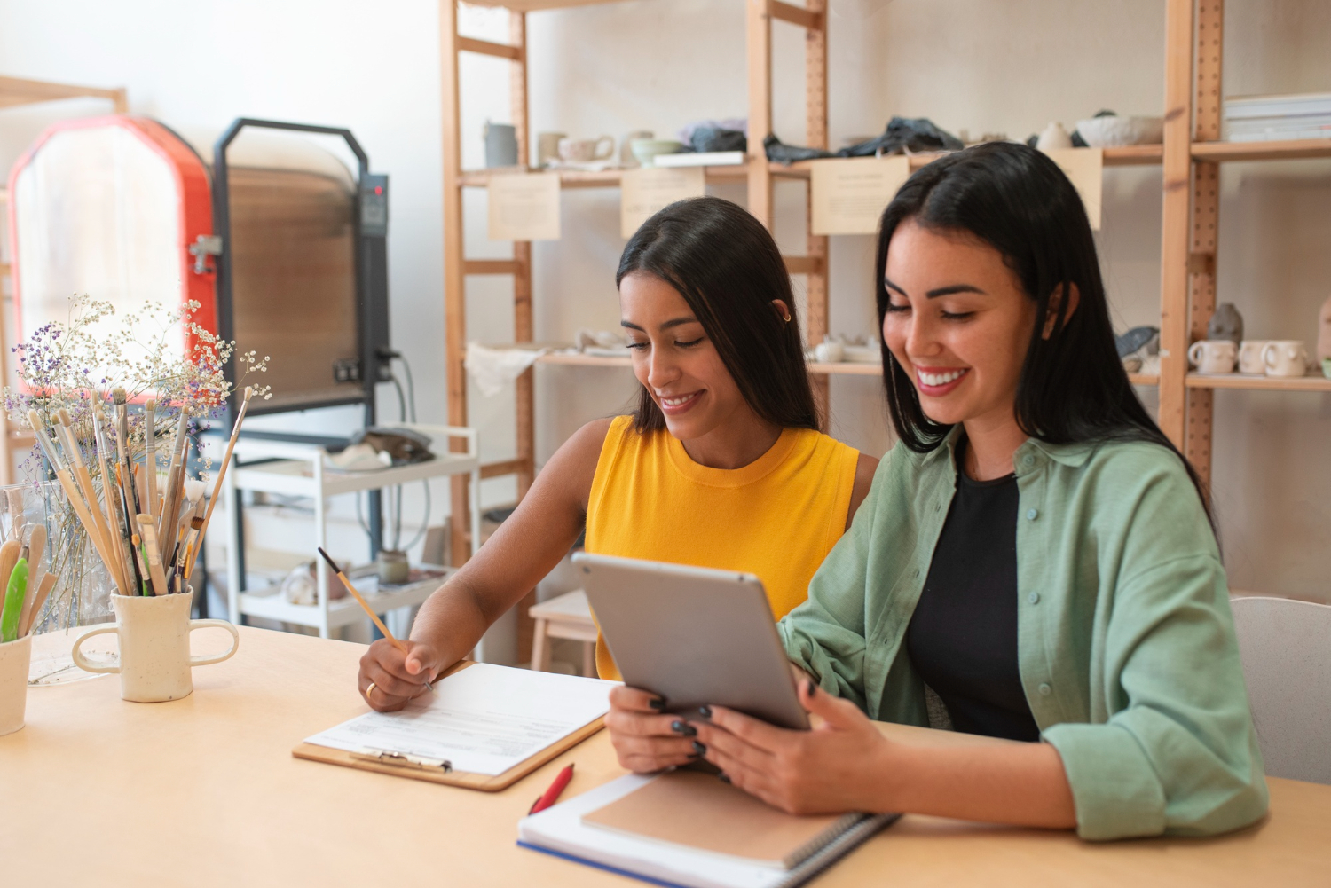 Dos mujeres sentadas en un escritorio trabajan juntas. De fondo se ve un anaquel. La imagen representa a dos emprendedoras.