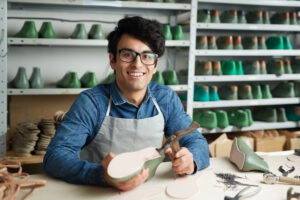 Un joven artesano sonriente con lentes y delantal gris trabaja en un taller de calzado. Sostiene una horma de zapato verde mientras posa frente a estanterías llenas de moldes y herramientas.
