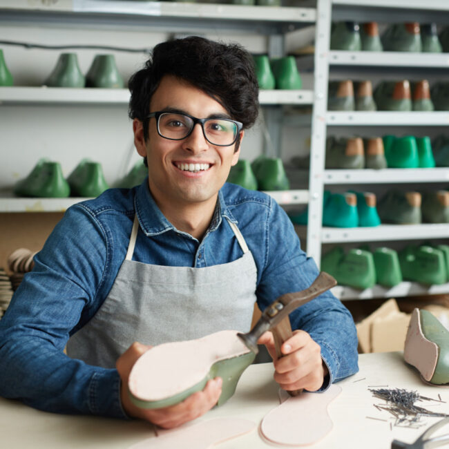 Un joven artesano sonriente con lentes y delantal gris trabaja en un taller de calzado. Sostiene una horma de zapato verde mientras posa frente a estanterías llenas de moldes y herramientas.