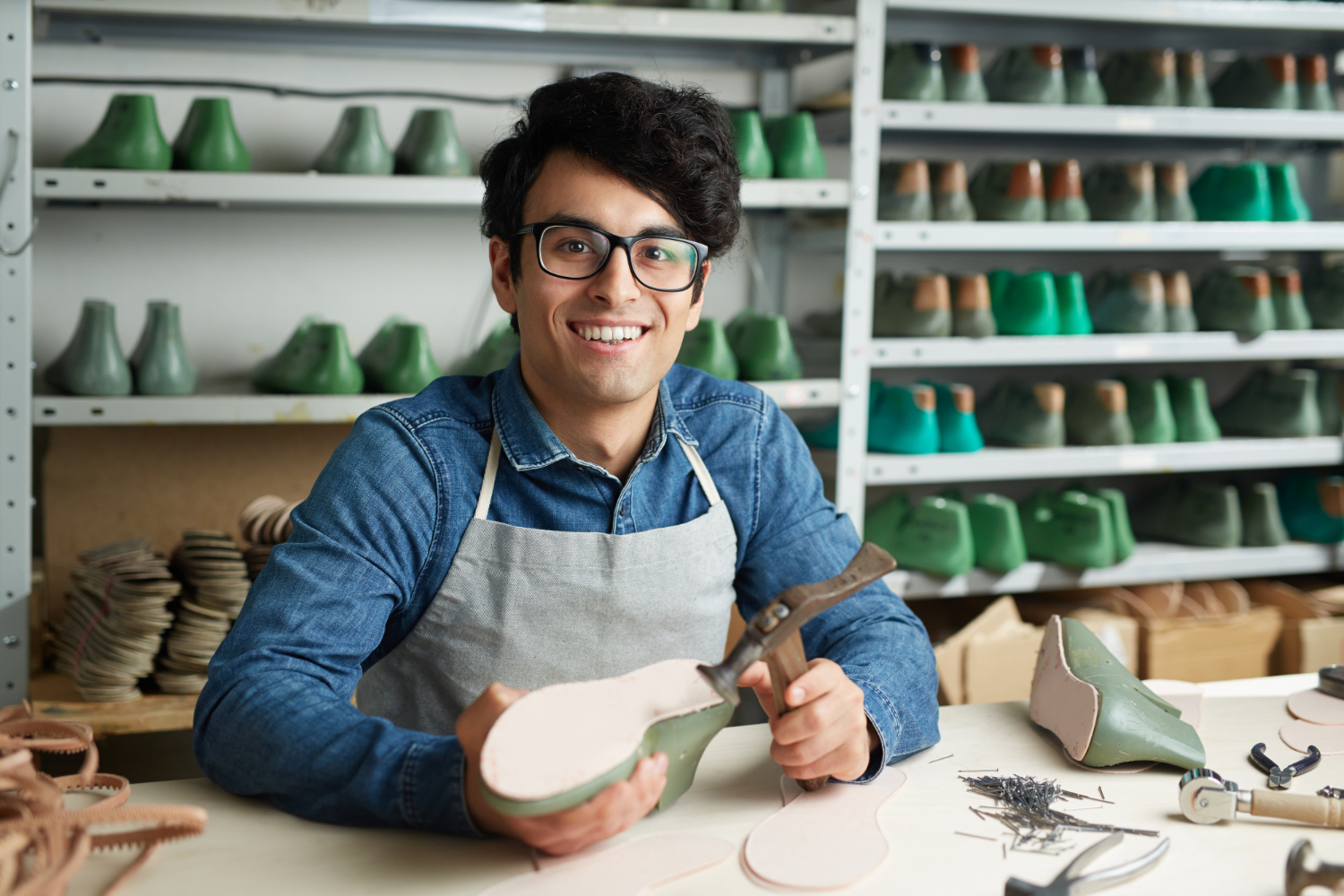 Un joven artesano sonriente con lentes y delantal gris trabaja en un taller de calzado. Sostiene una horma de zapato verde mientras posa frente a estanterías llenas de moldes y herramientas.